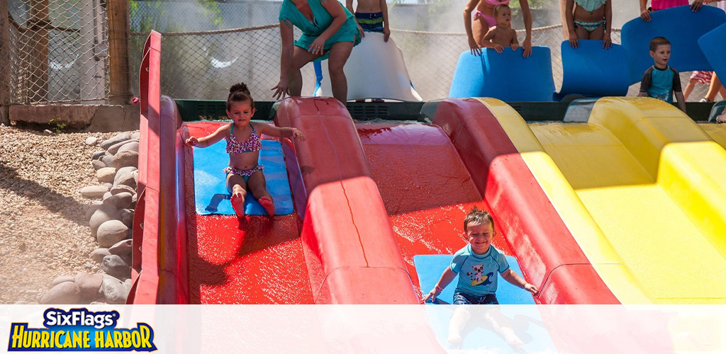 Image displays a vibrant water play area at Six Flags Hurricane Harbor. Children are enjoying a sunny day, sliding down colorful water slides in blue, red, and yellow with visible joy. The water sparkles, reflecting the daylight, as onlookers and waiting riders anticipate their turn. The environment suggests fun and excitement within a safe, family-friendly water park.