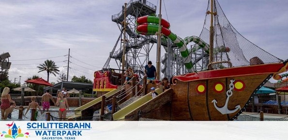 Image of Schlitterbahn Waterpark in Galveston, Texas. Visitors enjoy a sunny day with attractions including a pirate-themed playground and water slides in varying heights and sizes, featuring red, green, and white colors. Kid-friendly fun with a ship structure for climbing and playing.