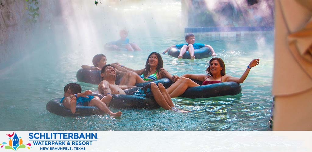 Several people are enjoying a lazy river ride at Schlitterbahn Waterpark and Resort in New Braunfels, Texas. They are floating on black inner tubes, some are laughing and talking, while mist and waterfalls enhance the tropical ambiance. The park's logo is visible in the bottom left corner.