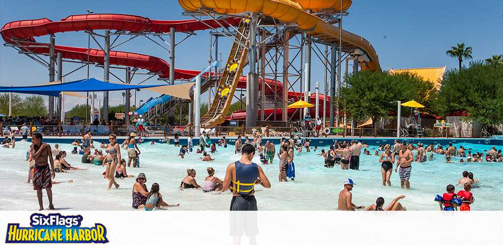 Visitors enjoy sunny weather at a bustling water park featuring a wave pool in the foreground and tall colorful water slides in the background. Yellow umbrellas and green palm trees dot the area with clear blue skies above.