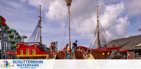 Image shows a sunny day at Schlitterbahn Waterpark in Galveston, Texas. Visitors enjoy a pirate-themed boat attraction with faux sails and a wooden plank. Water slides and park buildings can be seen in the background under a blue sky with scattered clouds.