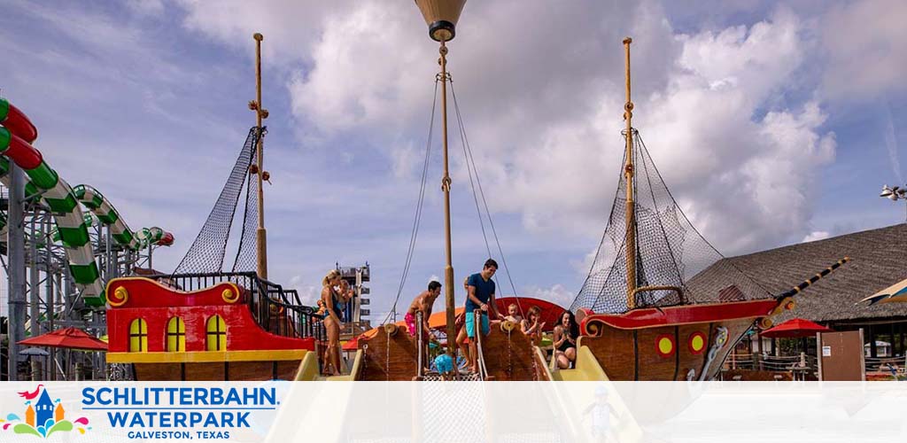 Visitors enjoy a sunny day at a waterpark in Galveston, Texas, with a focus on a colorful, pirate-themed play area. The image shows a bright red and yellow ship with nets and masts against a backdrop of waterslides and a blue sky with fluffy clouds.