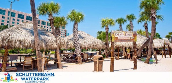 Image shows a sunny, sandy area with palm trees and thatched-roof cabanas at Schlitterbahn Waterpark in Galveston, Texas. The cabanas have tables and chairs for relaxation. A sign reading 'Cabana Beach' is visible in the distance.