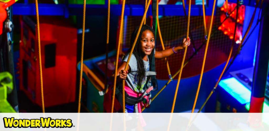 Child navigates a rope course indoors with safety harness, vibrant colors around.