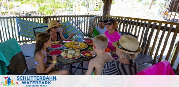 A group enjoys a meal under a shaded cabana at Schlitterbahn Waterpark, Galveston, Texas. There are palm trees in the background, signifying a tropical atmosphere. The table is set with various dishes, and the guests appear relaxed and engaged in conversation. The watermark indicates the location.