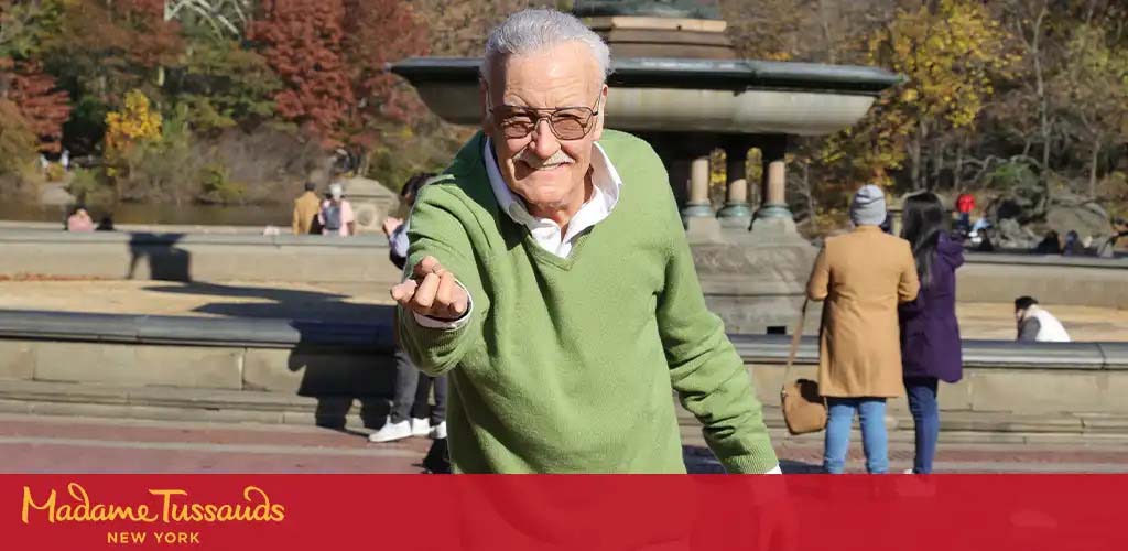 Image shows a wax figure of a smiling elderly gentleman pointing forward. He's wearing a white collared shirt, glasses, and a green cardigan. The background reveals an outdoor setting with people near a fountain, indicative of a public park. The bottom of the image has a red banner with the text 'Madame Tussauds New York'.