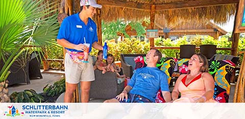 An employee at Schlitterbahn Waterpark & Resort, dressed in a blue uniform, interacts with two guests seated comfortably on a cabana lounge. The guests appear relaxed and engaged in conversation, with joyful expressions. Tropical decor and a thatched roof underscore the resort's leisurely ambiance.