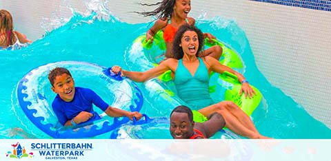 Two joyful kids and two excited adults are riding on bright blue and green inner tubes down a water slide at Schlitterbahn Waterpark. Splashes surround them as they enjoy the fun descent. The park's logo is displayed at the bottom.