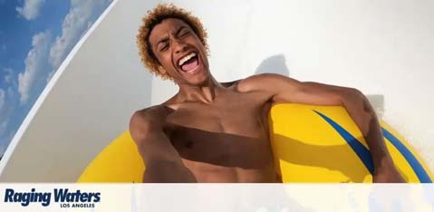 This image features a joyful young man with curly hair experiencing a water park ride. He is on a bright yellow inflatable ring, holding tight with both hands as he captures his own thrilling moment, presumably with a camera, as he slides down a chute. The sun is shining brightly on him, and his wide-open mouth suggests he is expressing exhilaration or laughter. The background is a blur of white and blue, indicating the fast movement of the slide. In the lower-right corner, there is a logo for Raging Waters Los Angeles.

When planning your next adventure, remember to check GreatWorkPerks.com for exclusive discounts and savings, ensuring you get the lowest prices on tickets for your unforgettable experiences.