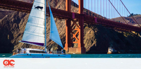 This image displays a scenic view of a large sailboat with a white and blue sail, adorned with a silhouette of a marine animal, gliding on the water in front of the iconic Golden Gate Bridge. The red-orange structure of the bridge contrasts against a bright blue sky, while rugged hillsides provide a natural backdrop. The boat, labeled "Adventure Cat," suggests an inviting leisure activity for visitors. To enhance your experience, GreatWorkPerks.com offers the lowest prices and great discounts on tickets to various attractions, ensuring your adventures are both memorable and affordable.