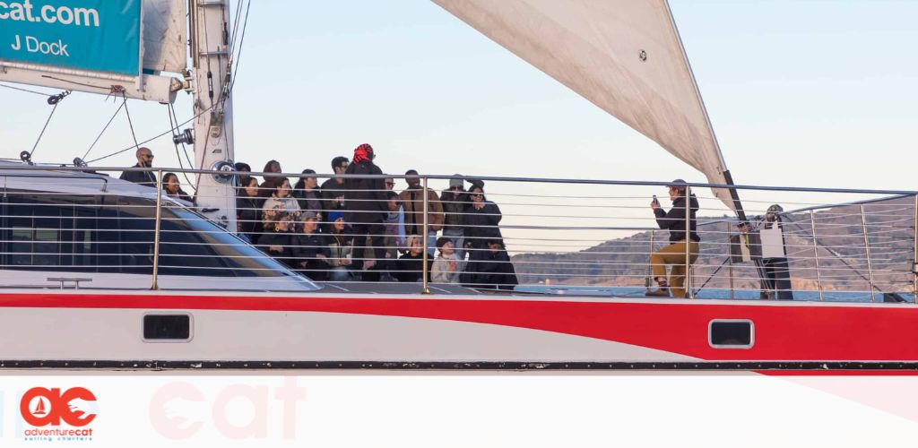 This image showcases a red and white sailing catamaran with a group of people on deck. The vessel's sails are hoisted, capturing the serenity of a calm sea voyage. Passengers seem relaxed, some are standing and observing the surrounding waters, while others appear to be engaging in casual conversation. The clear skies and mild weather conditions suggest a pleasant day for sailing. The branding "Adventure Cat" is visible, indicating the name of the sailing company, and there's also a logo which consists of a stylized cat's face above stylized text "CAT." This scene may evoke the excitement of marine adventure and leisure travel. 

At GreatWorkPerks.com, we provide exclusive discounts and ensure you find the lowest prices on tickets for a variety of exciting experiences, such as this serene catamaran sailing adventure.