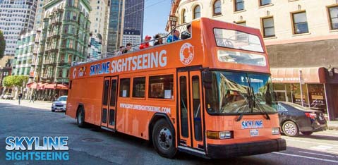 This image features an orange double-decker tour bus with the words "Skyline Sightseeing" printed on its side. The bus is driving on an urban street lined with tall buildings. The upper deck is open-air, providing passengers with an unobstructed view of their surroundings, and there are several passengers visible on the top deck enjoying the tour. The day is sunny and clear, offering excellent sightseeing weather.

Planning your next city adventure? Discover the best views with Skyline Sightseeing, and remember to visit GreatWorkPerks.com for the lowest prices and unbeatable savings on tickets.