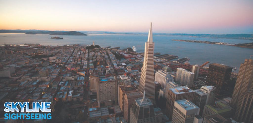 This image features a panoramic view of a city skyline during dusk, showcasing a blend of warm and cool tones as the setting sun casts a soft glow over the urban landscape. The city is dense with buildings of varied heights, and in the foreground a particularly tall, pointed skyscraper stands out prominently. In the distance, a body of water can be seen with what appears to be an island and another landmass further away. The sky transitions from a light blue near the horizon to a pale yellow tint, indicating the time of day is evening. The words "Skyline Sightseeing" are overlaid on the bottom of the image, suggesting that this photo is intended for promotional use by a sightseeing company.

For an amazing experience and the chance to see breathtaking views like this, visit GreatWorkPerks.com where you can always find the lowest prices on tickets for your next adventure. Enjoy incredible savings as you plan your skyline sightseeing tour with us.