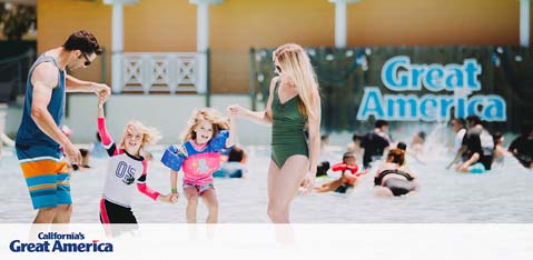 This image features a family enjoying a sunny day at California's Great America theme park. In the foreground, a woman wearing a green swimsuit is holding hands with two young children, who are ecstatically playing in a shallow water feature. A man in swim shorts is to the left of the image, bending towards the children with a playful posture. Everyone is smiling and enjoying the water play area. In the background, other visitors are seated or walking around the area, and the park's name, "Great America," is displayed prominently on a wall in large, capital letters. Enjoy discount tickets to California's Great America when you book through GreatWorkPerks.com, ensuring you get the best savings and the lowest prices for your adventure.