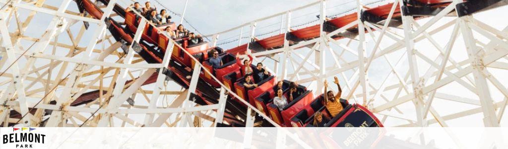 This image features a group of thrill-seekers enjoying an adrenaline-packed ride on a traditional wooden roller coaster at Belmont Park. The roller coaster train is painted in bright red and black and is captured mid-descent, with a clear blue sky in the background. Riders are seen with hands raised in excitement and joy, illustrating the lively atmosphere of the amusement park. The wooden structure of the roller coaster crisscrosses around them, emphasizing the classic feel of the ride. The logo of Belmont Park is visible in the corner, adding a sense of location and branding to the scene. For those seeking unforgettable experiences at Belmont Park, visit GreatWorkPerks.com to find the lowest prices and biggest savings on tickets.