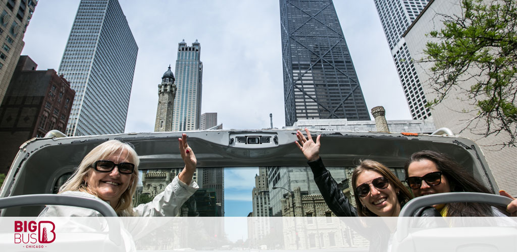 Image shows three people enjoying a sightseeing tour on a double-decker Big Bus in an urban environment. A joyful lady wearing sunglasses is visible in the front row, waving at the camera. Two more people are seated behind her; one is smiling and raising her hand high, while the other sits next to her. Tall modern skyscrapers fill the background under a bright sky. The Big Bus logo is in the bottom left corner.