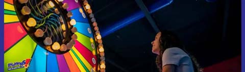 This image displays a colorful indoor amusement area where a person is engaging with a large, brightly lit, spinning wheel game. Visible joy and anticipation are evident on the individual's face as they look up at the spinning wheel, which features a spectrum of colors and numerous light bulbs that add excitement to the amusement atmosphere. The lighting within the space casts a warm glow, highlighting the dynamic nature of the game and the engrossed expression of the person enjoying the interactive entertainment on offer.

Discover the thrill of amusement with GreatWorkPerks.com, where outstanding entertainment meets incredible savings. Enjoy the lowest prices on tickets to your favorite attractions, ensuring your experiences are not just memorable, but also affordable.