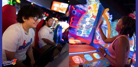 This image displays two individuals engaged in arcade games at a vibrant indoor gaming arena. To the left, a person with curly hair wearing glasses and a white t-shirt with blue lettering is seen intently focusing on a racing game, gripping a steering wheel attached to an arcade machine. Behind them, another individual is in the background, partially out of focus. To the right, a person with shoulder-length hair is interacting with a colorful, illuminated arcade game, pressing buttons on a panel with determination and enjoyment. The atmosphere appears lively and energetic, highlighted by the various bright lights and digital screens of the arcade setting. Remember, at GreatWorkPerks.com, we offer the thrill of unbeatable savings with discount tickets to the best entertainment venues for the lowest prices!