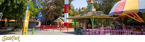 This image showcases a vibrant amusement park scene on a sunny day. The foreground features purple circular tables with matching chairs, inviting guests to relax and enjoy refreshments. To the right of the seating area, a whimsically designed carousel with a green roof and intricate embellishments hints at the classic park entertainment available to visitors. Lush trees can be seen in the background, providing a natural canopy and shade for the area. On the left, a bright blue and yellow ride structure adds a pop of color to the scene, suggesting more thrilling attractions nearby. The ground is paved, and the park appears well-maintained and ready for guests. Clearly visible in the bottom left corner is the name 'Castle Park' in fun and playful lettering, reinforcing the theme park's identity. At GreatWorkPerks.com, we're committed to offering our customers the best deals, ensuring you enjoy significant savings on tickets with the lowest prices available.