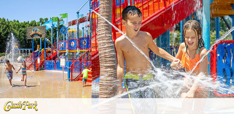 This image showcases an action-filled moment at a vibrant water park under a clear blue sky. The focal point is a water play structure with multiple levels of platforms and bright, colorful slides. A young boy and girl, the boy without a shirt and the girl in an orange swimsuit, are engaging in water play, smiling and splashing water on each other near the play structure. The boy appears to be pouring water from a small bucket, while the girl reacts with joy. Around them are additional water features, including water cannons and fountains, with sprays and jets creating a dynamic and refreshing scene. Palm trees are visible in the background, emphasizing the park's tropical theme. The logo of "Castle Park" is prominently displayed on the upper right-hand corner of the image, indicating the location of the depicted water play area. Remember, your day of fun in the sun is always within reach with our exclusive deals at GreatWorkPerks.com, where we promise the lowest prices on tickets and amazing savings for you and your family.