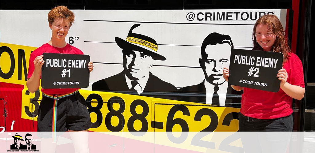 Two individuals stand smiling in front of a graphic that mimics a vintage mugshot backdrop, complete with height measurements. They hold signs saying 'Public Enemy #1' and 'Public Enemy #2'. The backdrop features black and white silhouettes of two men labeled '@CRIMETOURS'. The side of a red vehicle with the number 888-7622 and '@CRIMETOURS' is partially visible.