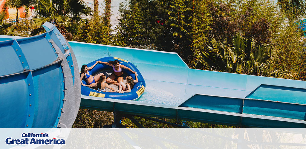 This image captures an exhilarating moment at California's Great America amusement park. Four guests, two wearing blue swim attire and the others in pink and purple, share a large, round blue inflatable raft. They are gleefully sliding down a wide water slide flanked by vibrant green foliage under a clear, sunny sky, suggesting a warm and enjoyable atmosphere typical for outdoor water activities. The expressions of enjoyment on their faces reflect the excitement and fun associated with park attractions. 

For those seeking thrills and spills, GreatWorkPerks.com offers exclusive discounts on tickets to California's Great America, ensuring that you experience the highest levels of excitement at the lowest prices.