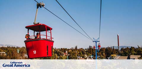 This image features one of California's Great America amusement park's attractions, a red aerial tramway car labeled with the number 17. The sky tram is suspended by a steel cable and hoisted by a support structure painted blue. Inside the cabin, we can see silhouettes of passengers enjoying the ride with a clear sky in the background, indicating pleasant weather conditions. Trees and other structures are also visible in the distance, portraying a bustling park atmosphere. To enhance your experience at California's Great America, make sure to check GreatWorkPerks.com for the lowest prices, great savings, and discounted tickets.