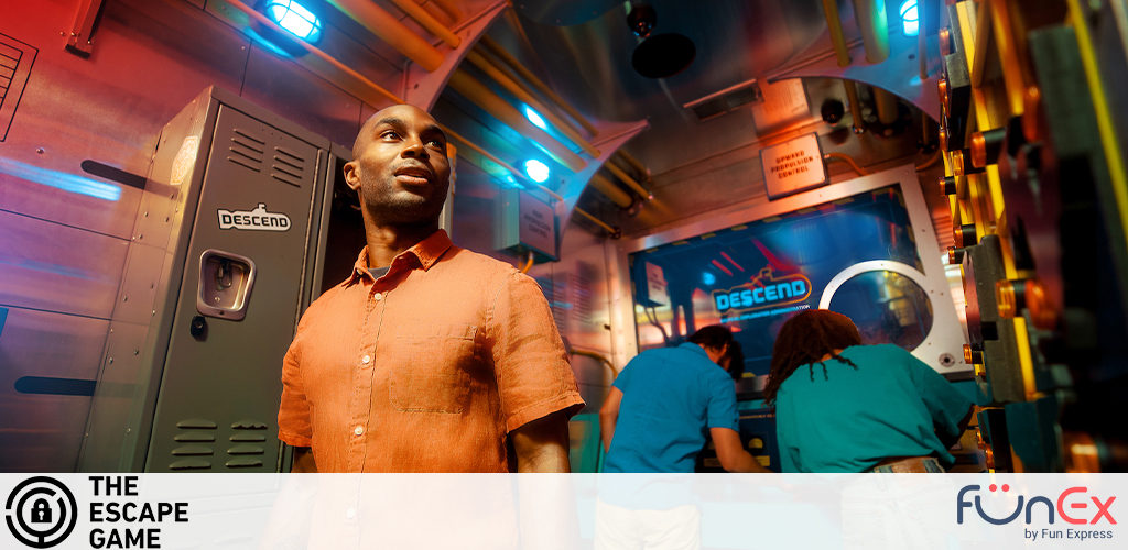 Image shows a brightly lit escape room with an industrial theme. In the foreground, a man in a burnt orange shirt looks around with a curious expression. In the background, three individuals in blue shirts are focused on deciphering clues around a centrally placed hatch-like door with the word DESCEND and neon blue lighting. Logos indicating 'The Escape Game' and 'FunEx by Fun Express' are visible.