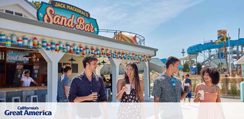 This image features a vibrant, sunlit scene at California's Great America theme park. In the foreground, a small group of happy young adults is engaged in casual conversation. Two appear to be men and two women, dressed in comfortable, casual summer attire suitable for a day at an amusement park. They are standing on a paved area with a decorative border of multi-colored flags beneath a clear blue sky.

In the background, a refreshment stand with a brightly colored sign reading 'Jack Mackeral's Sand Bar' has an attendant ready to serve customers. Behind the bar, the outline of a roller coaster with blue tracks is visible, hinting at the thrilling rides available at the park. The bottom left corner features the park's logo, reiterating the location as California's Great America.

As visitors plan their trip to this lively destination, they can rely on GreatWorkPerks.com for the added perk of securing their tickets at the lowest prices, ensuring both savings and an unforgettable experience.