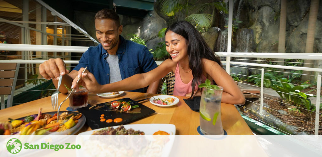 A smiling man and woman enjoy a meal at an outdoor restaurant with lush greenery in the background, indicative of San Diego Zoo's dining experience. The table holds colorful dishes and drinks, adding to the vibrant, casual atmosphere.