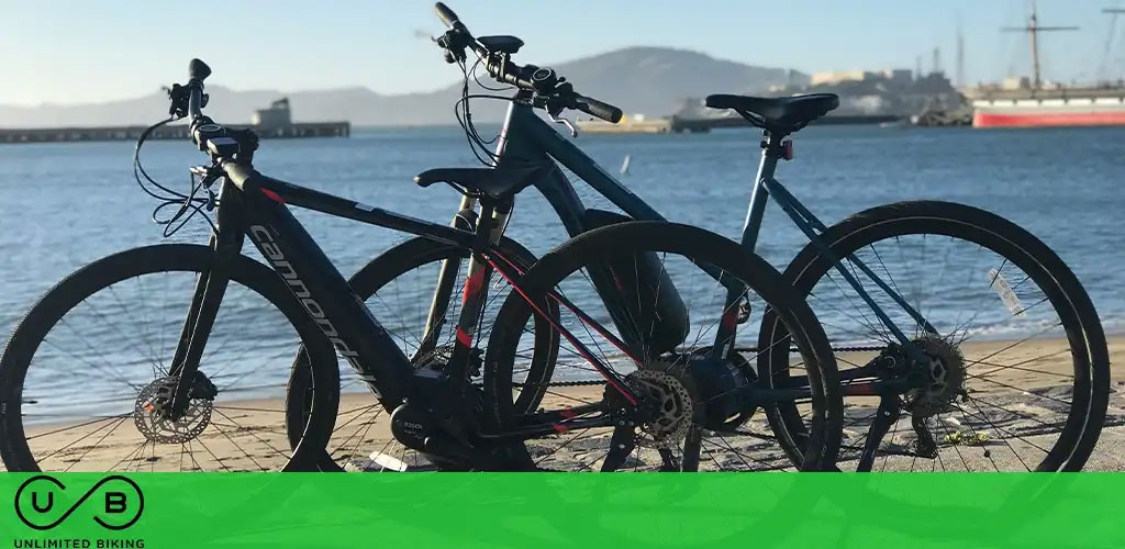 Two bicycles by a lakeside with a clear sky, a distant hill, and a pier in the background.