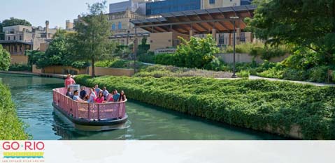 Image of a vibrant, scenic waterway with people enjoying a guided boat tour. The boat is pink and blue with GO RIO printed on the side, cruising along green lush banks under the clear blue sky. Modern buildings overlook the calm river, adding to the urban oasis vibe.