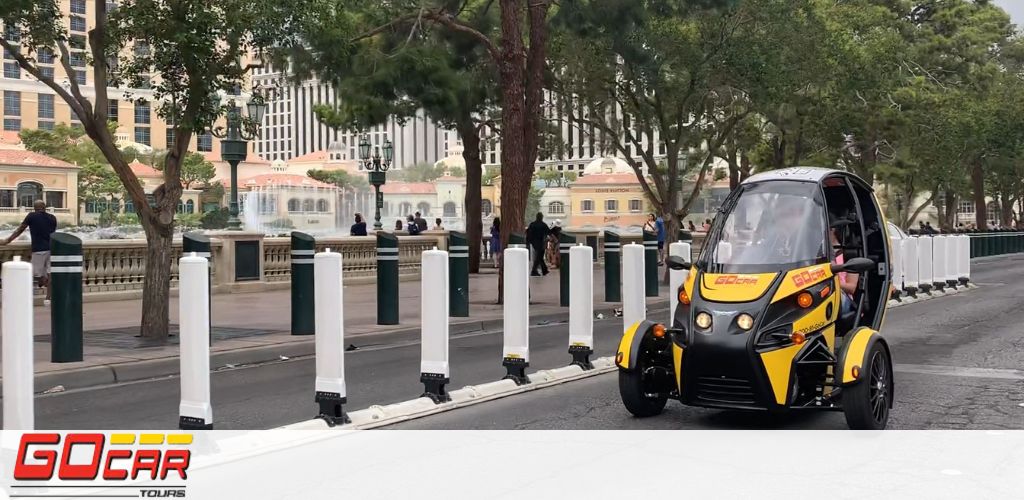 Image shows a yellow and black three-wheeled GoCar driving on a city street lined with trees and bollards. In the background, there are people walking and a fountain, hinting at an urban recreational area. The GoCar logo is prominent on the vehicle and the image.