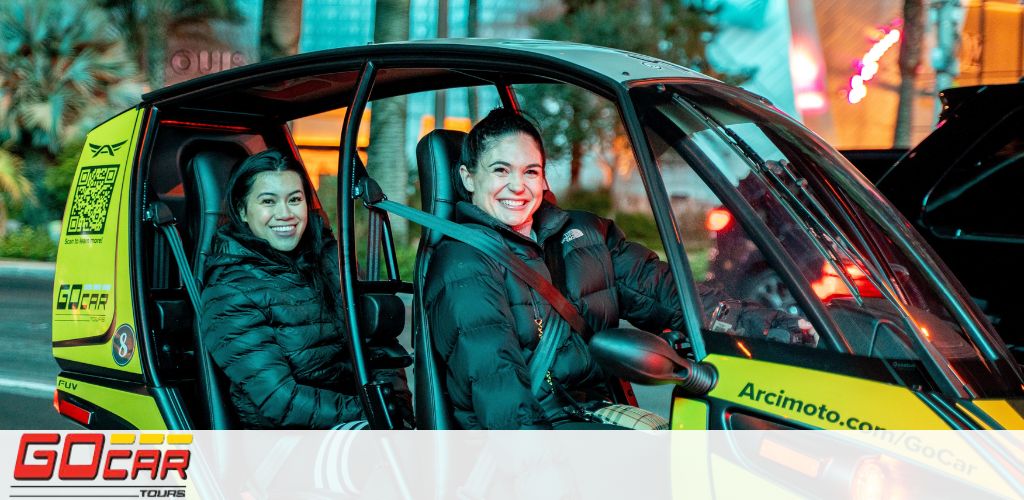 Two women are smiling from inside a yellow and black three-wheeled GoCar during what appears to be dusk, based on the lighting. The vehicle's branding emphasizes city tours, with decals and a QR code on the side. The urban backdrop hints at a lively atmosphere with blurred city lights.