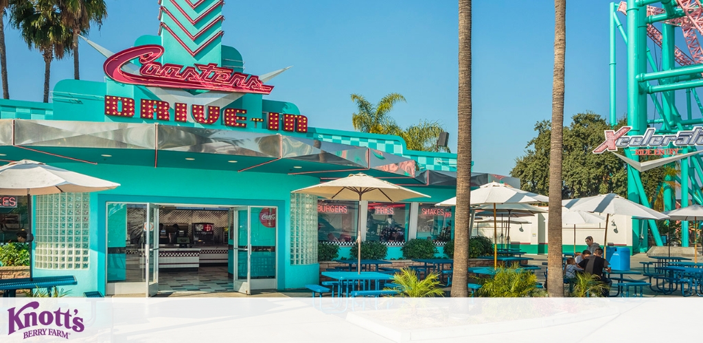 Image displays a vibrant retro-style diner with turquoise and pink colors, showing outdoor seating under white umbrellas. A rollercoaster is visible in the background, indicating the setting is in an amusement park. Clear skies and palm trees enhance the sunny ambiance.