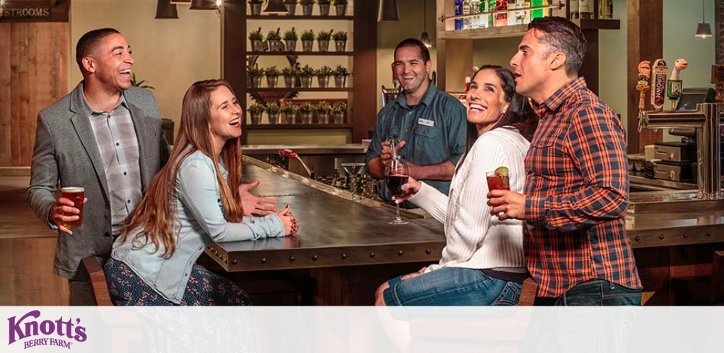 Image Description: This is a lively scene set at a bar within Knott's Berry Farm. There are two couples at the bar counter displaying expressions of enjoyment and camaraderie. On the left, a man in a gray blazer and grey checkered shirt holds a beer while smiling broadly beside a woman in a light blue cardigan and dark top, who is laughing and leaning slightly towards the man. On the right side, another couple is engaged in animated conversation with the woman wearing a white knit sweater and the man in a checked shirt, both holding wine glasses. Behind the bar, a smiling bartender in a blue uniform is interacting with the guests. The back wall behind the counter is adorned with neatly arranged shelves stocked with various bottles and glassware, adding to the warm and inviting atmosphere of the venue. The Knott's Berry Farm logo is visible in the left lower corner of the image.

Remember to check out GreatWorkPerks.com for unbeatable savings on tickets to Knott's Berry Farm, ensuring you get the lowest prices for your next fun-filled adventure!