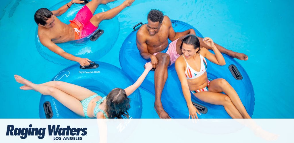 Four people enjoy a sunny day at Raging Waters Los Angeles, floating on blue inflatable rings in a clear, blue water park pool, conveying fun and relaxation.