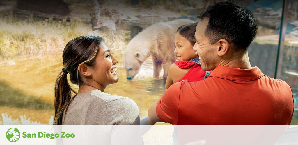 A family enjoys a day out at the San Diego Zoo. A smiling woman looks over her shoulder, while a man carries a young child on his back, both admiring a large polar bear on display. The zoo's logo is visible in the corner. Warm lighting complements the scene of bonding and discovery.