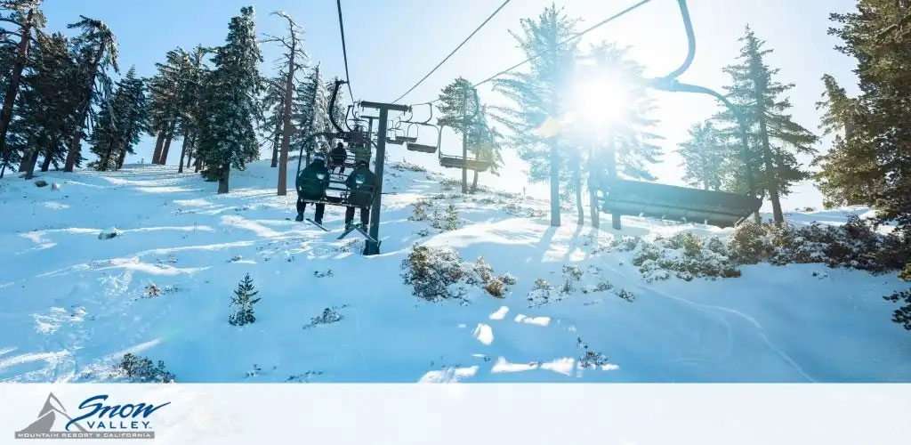 This image displays a serene winter scene at Snow Valley Mountain Resort in California, where a ski lift carries two visitors above a glimmering blanket of snow. Majestic pine trees dot the landscape, partially covered in snow, standing tall against a bright blue sky. The sun shines from the right side of the frame, casting its warm glow and creating sunbursts that peek through the trees. The ski lift travels upwards to the left, suggesting an ascent toward higher slopes. The Snow Valley logo is visible in the lower left corner, signifying the resort's branding. 

Experience exhilarating high-altitude adventures and savor the savings; at GreatWorkPerks.com, you'll always find the lowest prices on tickets to make your snowy escapades even more enjoyable!