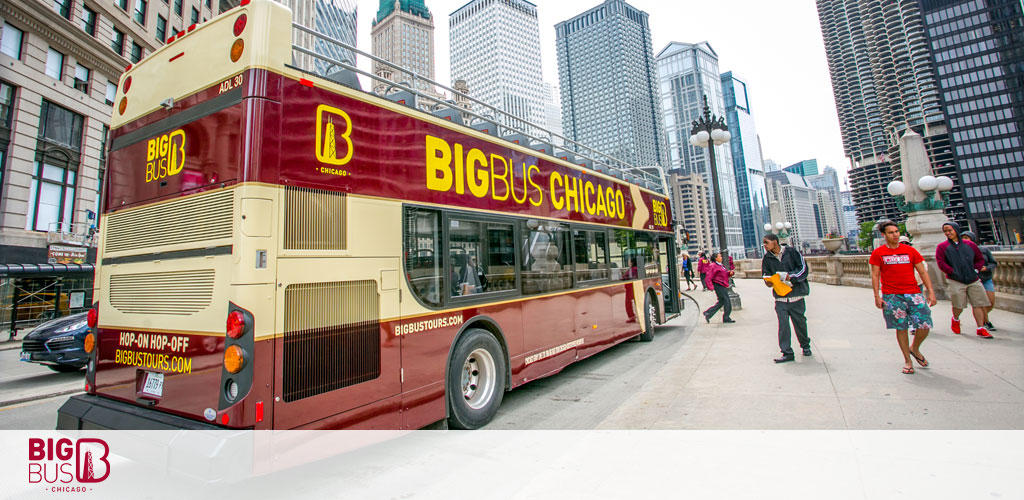A red Big Bus city tour bus in Chicago with a few people walking on the sidewalk and the city's skyscrapers in the background. The bus has signage that reads 'Hop-on Hop-off' and the company's website.