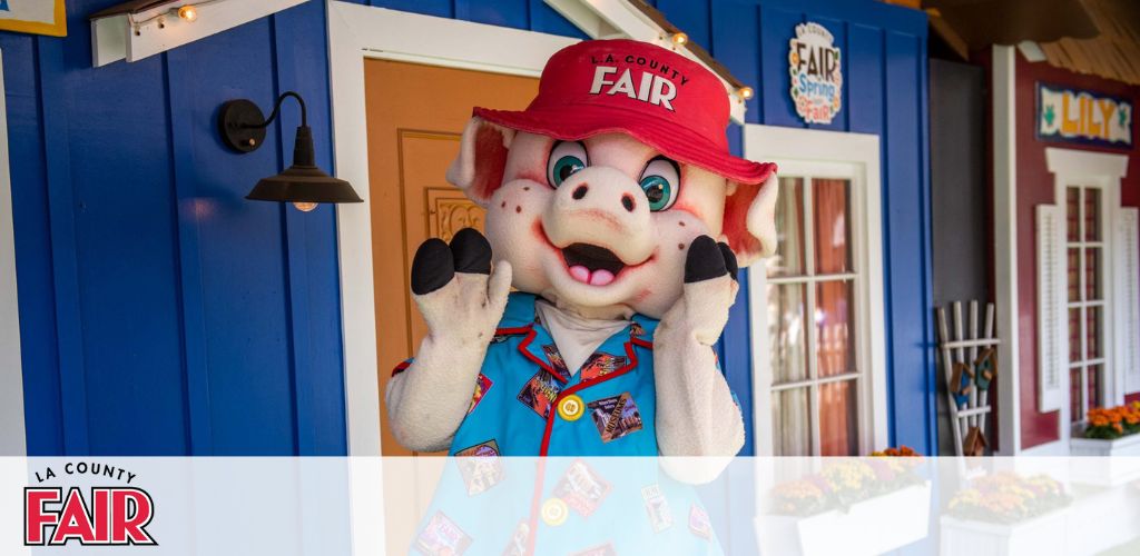 This image features a vibrant scene from the LA County Fair. In the foreground, a cheerful costumed character resembling a pig is standing and waving. The character is sporting a red baseball cap labeled "FAIR" and a colorful shirt adorned with various fair-themed patches. Behind the character, a quaint blue building facade with orange flowers at the base offers a picturesque backdrop, complete with a "LILY" sign on a neighboring red structure. The warm and inviting ambiance suggests the fun and excitement typical at county fairs.

At GreatWorkPerks.com, we're committed to making your visit to the fair an unforgettable experience with the added benefit of savings; enjoy the lowest prices on tickets when you plan your next adventure with us!