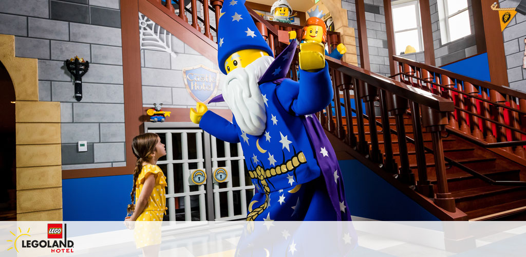 Image Description:
This vibrant photograph captures an enchanting moment inside the LEGOLAND Hotel. In the foreground of the image, a child dressed in a yellow dress with polka dots is engaged in an interaction with a life-size LEGO mascot character, the wizard, who is clothed in an elaborate blue-and-yellow costume adorned with stars. The wizard is extending a friendly hand towards the child. Both are standing in a hallway characterized by LEGO aesthetic details, including gray brick wallpaper and decorations that convey a medieval castle theme. To the right, a wooden staircase with a rich brown hue leads to an upper level. In the background, we can also spot two LEGO figures perched playfully on the railing of the staircase—one appearing to be a knight and the other a construction worker. The bright, whimsical setting evokes a sense of adventure and creativity synonymous with the LEGO brand.

End Statement:
Explore the magical world of LEGOLAND and create lasting memories with your family! Don't forget to check GreatWorkPerks.com for exclusive discounts and the lowest prices on tickets to your favorite attractions.