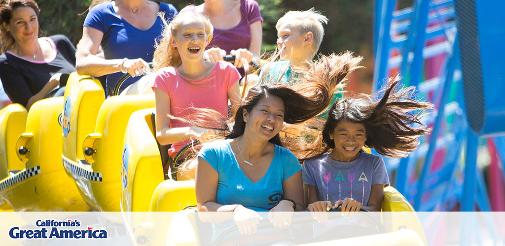 This image features a diverse group of people enjoying a thrilling ride at California's Great America amusement park. In the foreground, two young girls with vibrant smiles are seated in a bright yellow roller coaster car, their hair wildly flying behind them, suggesting the car is moving at high speed. The sun shines brightly, illuminating the scene with a warm glow and highlighting the excitement and joy on the riders' faces. Trees form a green backdrop to the image, signifying an outdoor setting. The amusement park's logo is visible in the upper left corner, emphasizing the venue.

Experience the thrill of California's Great America and save on your next adventure with us at GreatWorkPerks.com, where we offer the lowest prices on tickets, ensuring your day is not only exhilarating but also easy on your wallet!