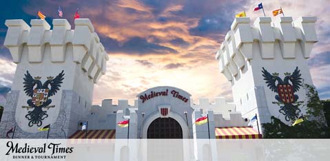 Image shows the facade of Medieval Times Dinner & Tournament, featuring castle-like architecture with twin towers against a sunset sky. Banners adorn the building, emblazoned with medieval emblems. The venue's name is prominently displayed.