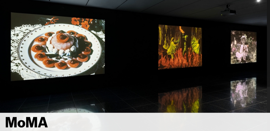 The image shows an interior view of a dimly lit art gallery with three large, colorful projections on the walls. On the left, a photo of food presentation on a plate. The center and right projections depict abstract, vibrant art and a vintage photo with a figure, respectively. The reflective floor enhances visual depth, and the MoMA logo is seen at the bottom center.