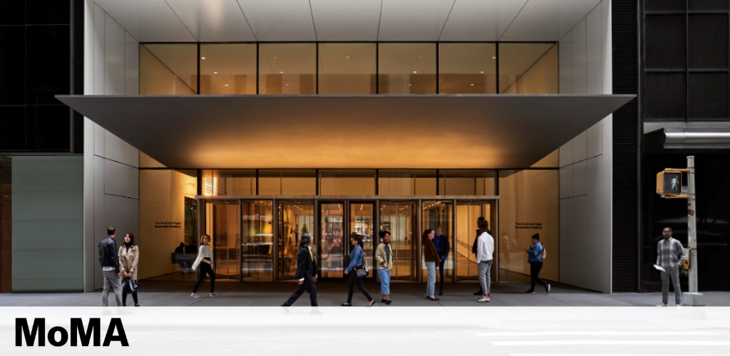 The image shows the modern entrance of the MoMA with its name displayed prominently at the bottom. Warm lighting emanates from the canopy above the glass doors. People are seen walking by and entering the building, illustrating a lively and accessible venue.