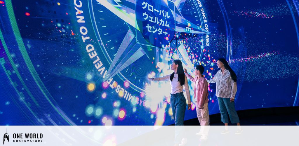 Three individuals stand in front of a large, illuminated circular display with dynamic graphics and text highlighting the New York City skyline. They appear engaged, pointing at the vibrant visual elements that represent the city's energy. The One World Observatory logo is in the corner, suggesting this is an exhibit within the attraction. The scene is lively with a blue and pink light scheme.