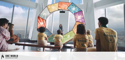 A group of visitors is seated indoors facing a presenter who stands near a display shaped like a ring with various screens. The room has large windows offering a view of the cityscape far below. The attendees appear engaged, with some wearing shirts that read  One World Observatory. 