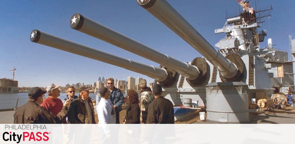 Group of people on a sunny day taking a tour around the large deck guns of a naval warship with the city skyline in the distance. The words 'Philadelphia CityPASS' are displayed as a promotional banner.