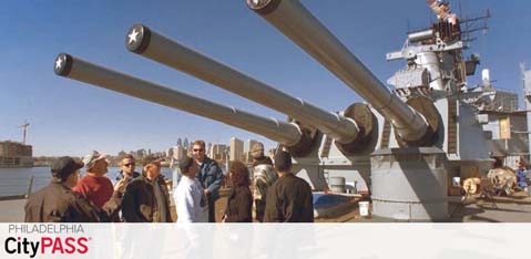 Image shows a group of visitors on a navy ship's deck, standing near the large naval gun barrels under a clear sky. The caption 'Philadelphia CityPASS' suggests a tourist experience.